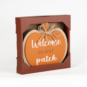 An orange heart-shaped wooden sign with white script text reading 'Welcome to our patch' and a jute twine bow, displayed inside a brown cardboard gift box on a white background.