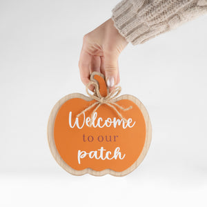 A hand wearing a beige knit sweater holds a pumpkin-shaped wooden sign that reads 'Welcome to our patch' in white and brown lettering against an orange background, set against a clean white studio backdrop.