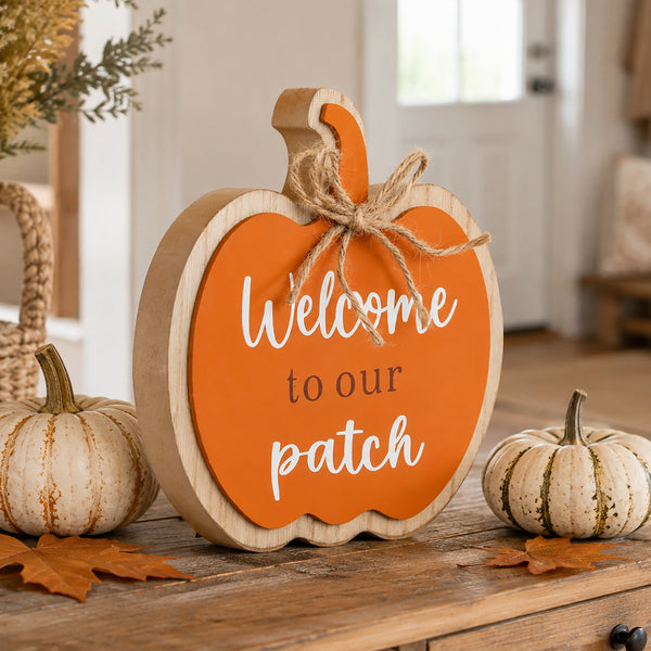 A scalloped wooden pumpkin sign with an orange front and natural wood sides, reading “Welcome to our patch” in white script and serif text, tied with a jute bow. It sits on a wooden table in a warm, decorated home setting with small pumpkins and autumn leaves.
