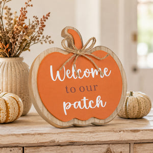 A scalloped pumpkin-shaped wooden sign with an orange face, white script reading 'Welcome to our patch,' and a rustic twine bow on the stem, styled on a wooden surface with fall decor in a softly lit indoor setting.