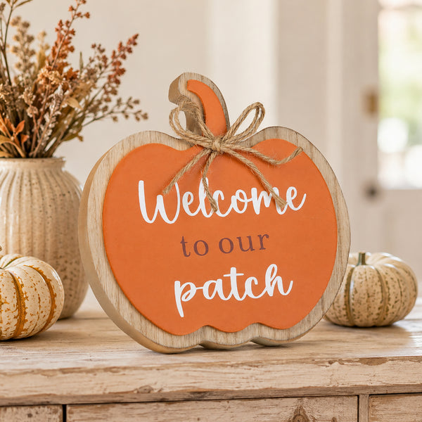 A scalloped pumpkin-shaped wooden sign with an orange face, white script reading 'Welcome to our patch,' and a rustic twine bow on the stem, styled on a wooden surface with fall decor in a softly lit indoor setting.