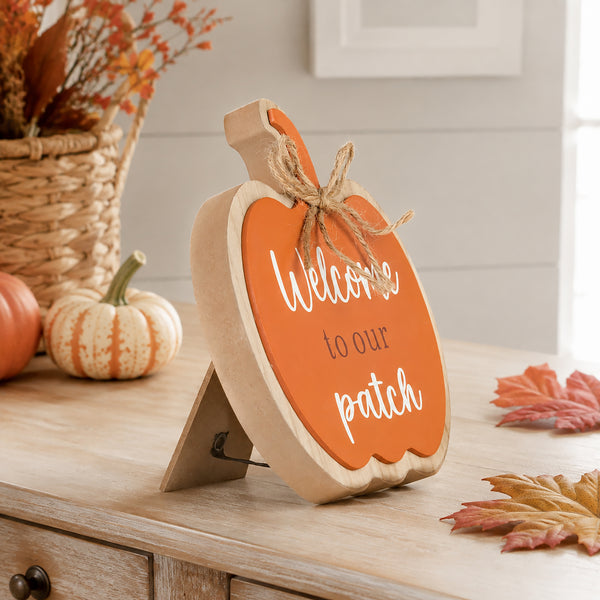 A decorative wooden pumpkin sign with a natural wood frame and handle, orange front panel, white script text reading 'Welcome to our patch,' and a jute bow, displayed on a wooden table in a fall-themed setting.