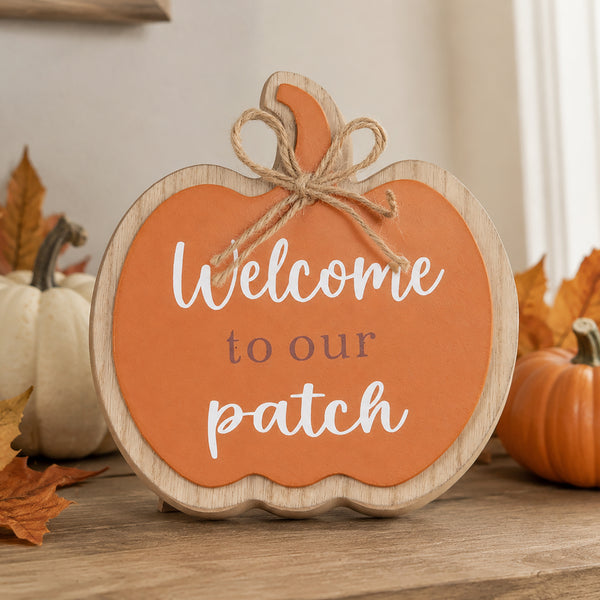 A pumpkin-shaped wooden sign with a scalloped edge, featuring an orange center with the phrase 'Welcome to our patch' in white and gray lettering. The sign has a natural wood border, a jute twine bow at the top, and is styled on a wooden surface with autumn leaves and small pumpkins in the background.
