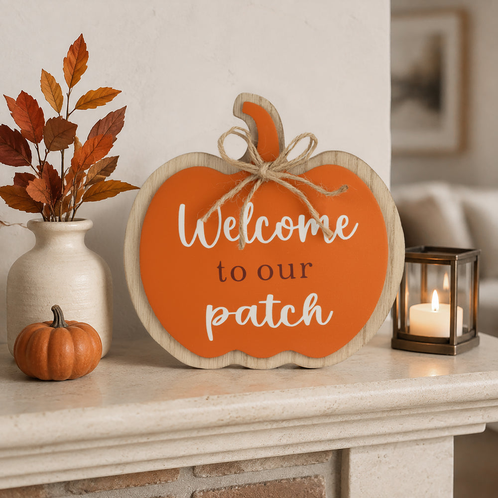 An autumn-themed wooden pumpkin sign with the text 'Welcome to our patch' in white and black lettering, styled on a fireplace mantel with a small orange pumpkin, a ceramic vase with fall leaves, and a lit candle.