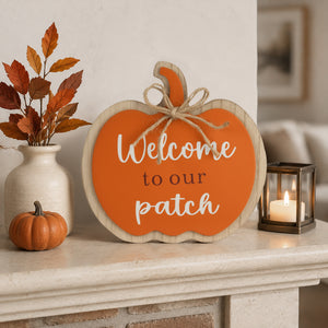 An autumn-themed wooden pumpkin sign with the text 'Welcome to our patch' in white and black lettering, styled on a fireplace mantel with a small orange pumpkin, a ceramic vase with fall leaves, and a lit candle.