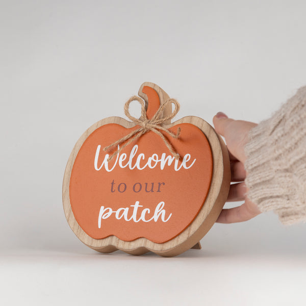 A hand wearing a beige knit sweater holds a wooden pumpkin-shaped sign with a terracotta orange face, white script text reading 'Welcome to our patch,' and a small jute bow on a light gray background.