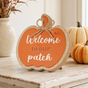 A scalloped wooden pumpkin sign with an orange face and natural wood border, reading 'Welcome to our patch' in white and brown lettering, styled on a rustic white table with pumpkins and a vase in the background.