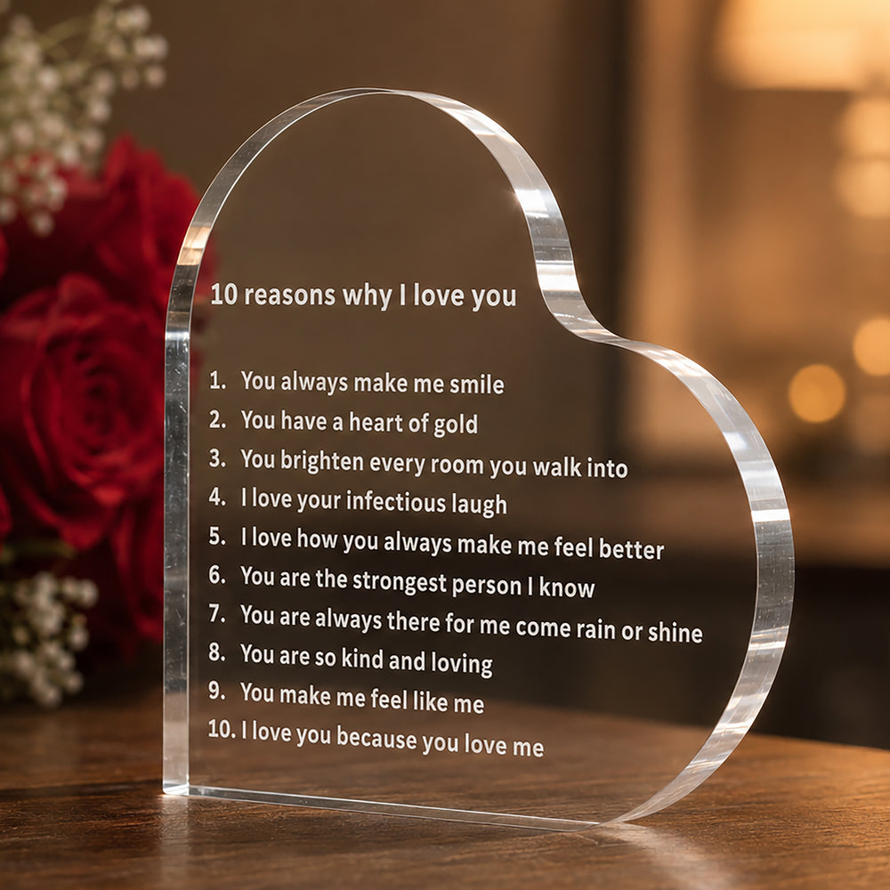 A clear acrylic heart-shaped plaque engraved with the text '10 reasons why I love you' and a poem, displayed on a wooden surface with a blurred background of red roses and warm bokeh lights.