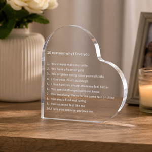 A heart-shaped clear acrylic plaque engraved with '10 reasons why I love you' sits on a wooden table in a softly lit home setting, with a vase of white flowers and a candle blurred in the background.