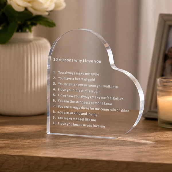A heart-shaped clear acrylic plaque engraved with '10 reasons why I love you' sits on a wooden table in a softly lit home setting, with a vase of white flowers and a candle blurred in the background.