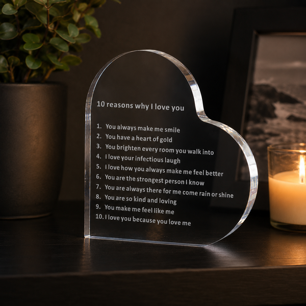 A heart-shaped clear acrylic plaque engraved with the text '10 reasons why i love you' sits on a dark wooden table. The background is dimly lit with a potted plant on the left and a lit candle in a glass jar on the right.