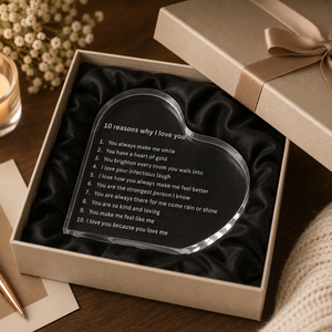 A clear acrylic heart-shaped plaque engraved with '10 reasons why I love you' sits inside an open beige gift box lined with black satin. The box is wrapped with a beige ribbon and styled on a wooden surface with a candle, dried flowers, and a pen nearby.