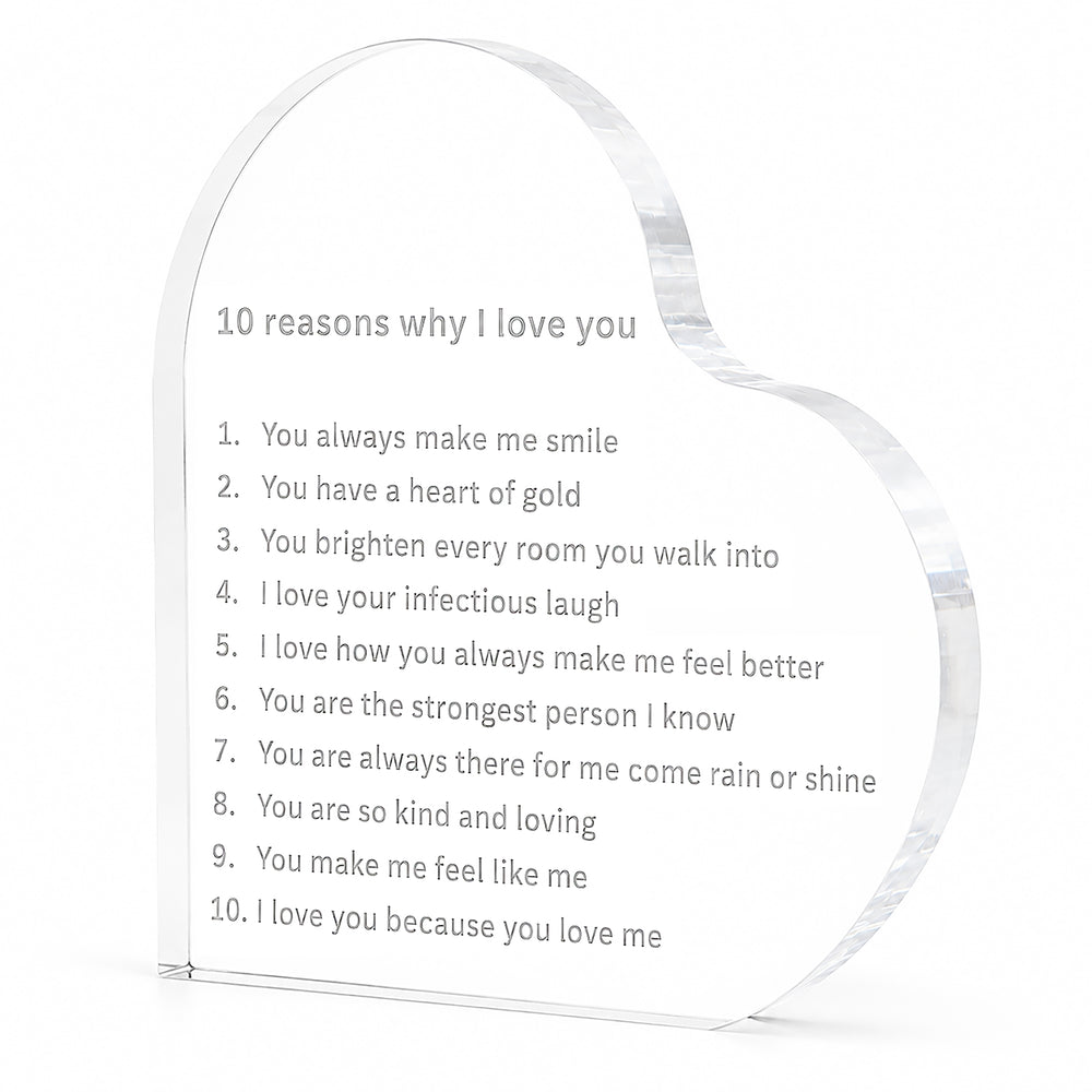 A clear acrylic heart-shaped plaque engraved with the text '10 reasons why I love you' and a poem, displayed on a wooden surface with a blurred background of red roses and warm bokeh lights.
