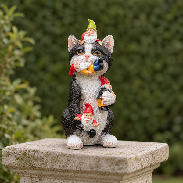 A whimsical garden statue of a black-and-white cat sitting on a stone pedestal, holding several small garden gnomes and a green leaf in its paws. The background is a soft-focus green garden hedge.