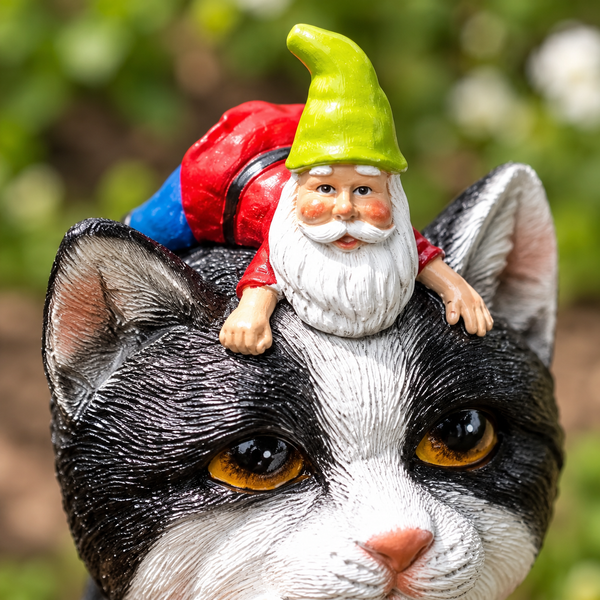A close-up of a black-and-white cat head garden statue with a small gnome figurine sitting on its forehead. The gnome wears a bright green hat, red jacket, and white beard.