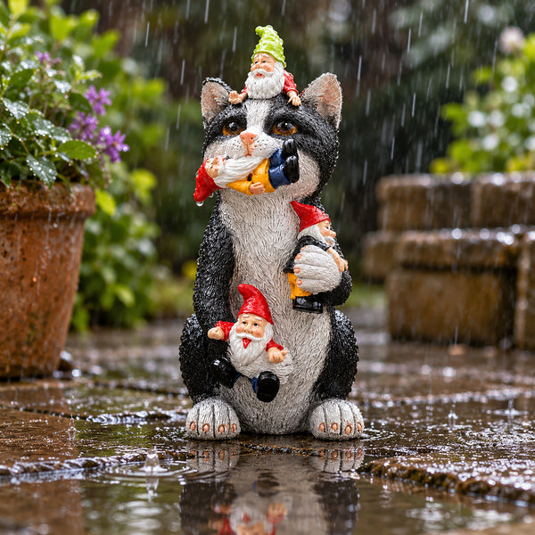 A black and white cat garden statue with three small garden gnomes, shown outdoors in the rain on a wet stone surface with blurred plants in the background.