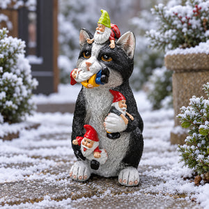 A decorative garden statue of a black-and-white cat carrying three small gnome figures, photographed outdoors on a snow-dusted patio with shrubs and a stone planter in the background.