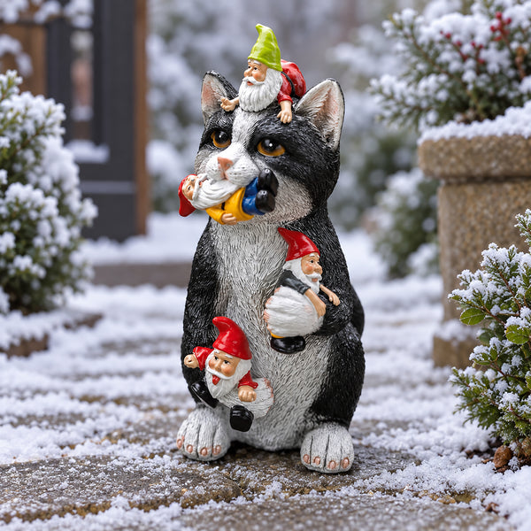 A decorative garden statue of a black-and-white cat carrying three small gnome figures, photographed outdoors on a snow-dusted patio with shrubs and a stone planter in the background.