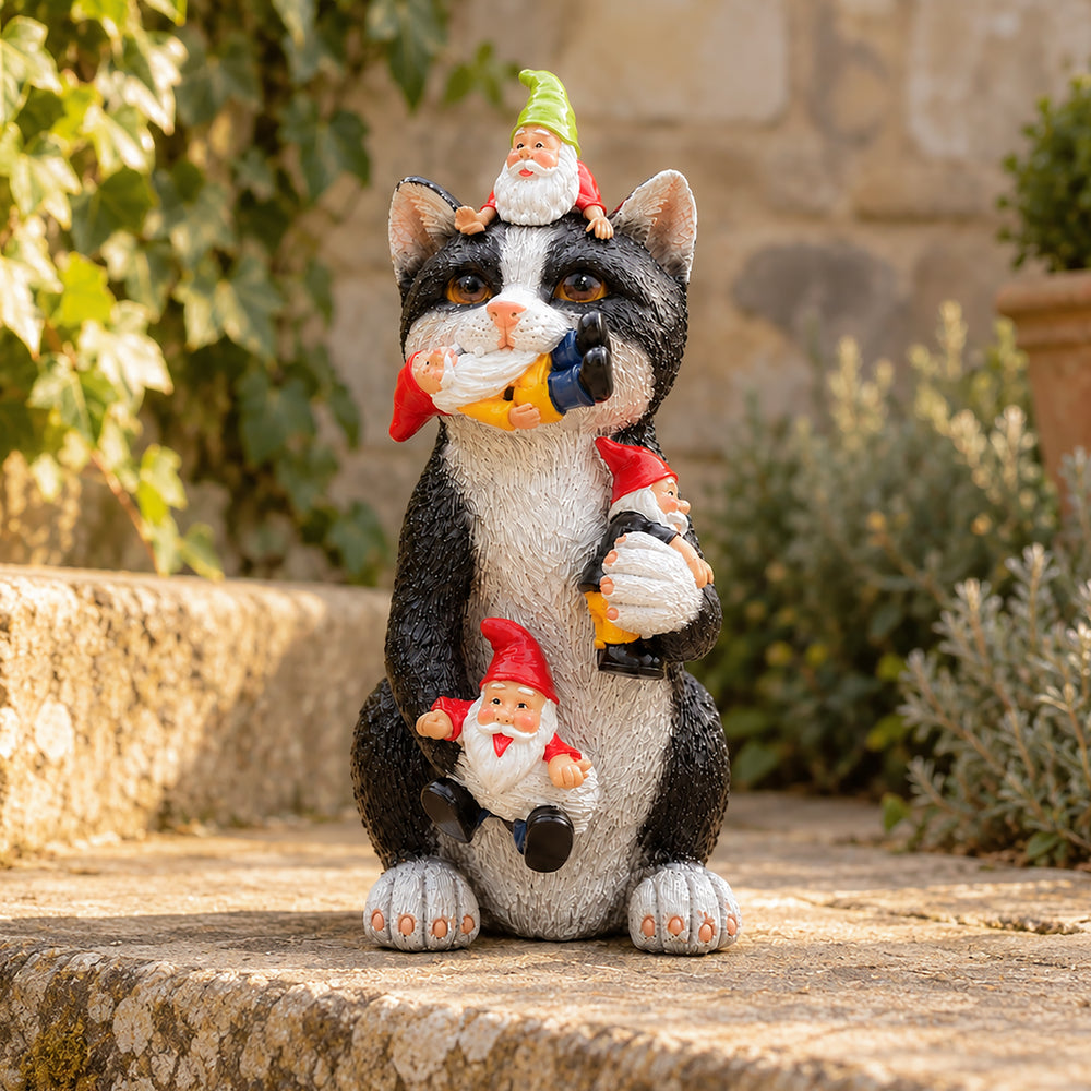 A black and white cat garden statue holding three small gnome figures in its paws, set outdoors on a stone ledge with green plants and a stone wall in the background.