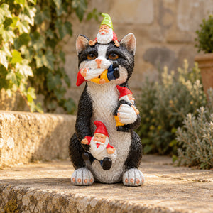 A black and white cat garden statue holding three small gnome figures in its paws, set outdoors on a stone ledge with green plants and a stone wall in the background.
