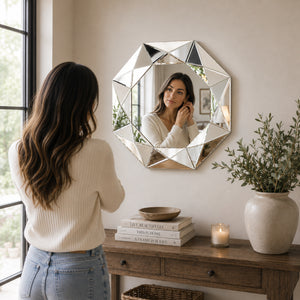 A woman stands with her back to the camera, looking at her reflection in a large geometric octagonal wall mirror hanging above a wooden console table in a softly lit room.
