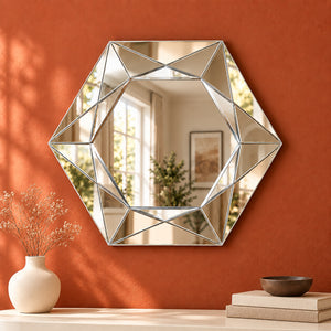 A large hexagonal wall mirror with a geometric faceted silver frame, mounted on a textured terracotta orange wall above a console table styled with a vase of dried flowers and books.