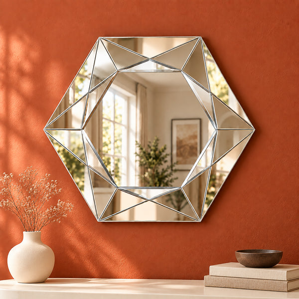 A large hexagonal wall mirror with a geometric faceted silver frame, mounted on a textured terracotta orange wall above a console table styled with a vase of dried flowers and books.