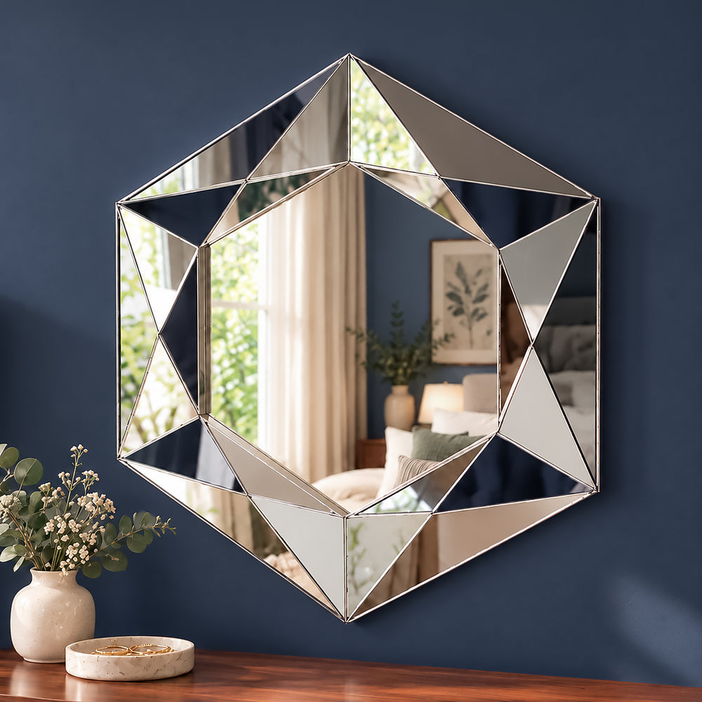 A large geometric hexagonal wall mirror with a silver frame hangs on a dark blue wall above a wooden table with a vase of flowers and a small dish. The mirror reflects a softly lit room interior.