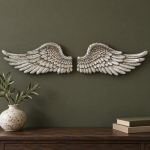 A pair of distressed silver angel wings wall decor mounted on a dark olive green wall above a wooden console table with a ceramic vase and books.