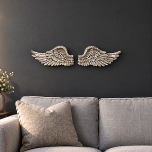 A pair of metallic silver angel wings wall decor mounted on a dark charcoal wall above a modern gray fabric sofa with a neutral throw pillow.