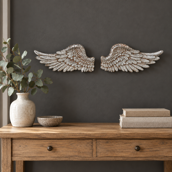 A pair of distressed silver angel wings wall art mounted on a dark gray wall above a wooden console table styled with a vase, bowl, and books.
