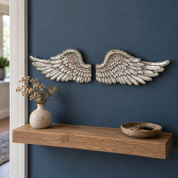 A pair of large textured silver angel wings wall decor mounted on a dark blue wall above a wooden floating shelf with a small vase and bowl.
