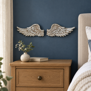 A pair of textured, metallic silver angel wings wall art mounted on a dark blue wall above a wooden nightstand with a vase and book.