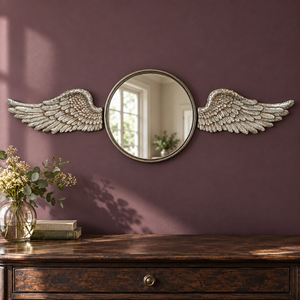 An ornate round wall mirror with decorative silver angel wings on each side, mounted on a dark purple wall above a wooden console table with a vase of flowers.