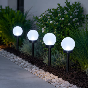 Four glowing spherical solar garden lights mounted on black stakes along a stone walkway in a landscaped garden at night.