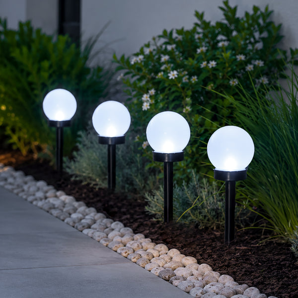 Four glowing spherical solar garden lights mounted on black stakes along a stone walkway in a landscaped garden at night.