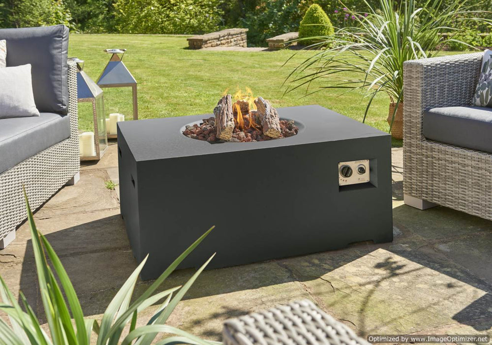 An outdoor fire pit with a black square composite body, featuring ceramic logs and lava stones inside, set on a patio with outdoor furniture around it.