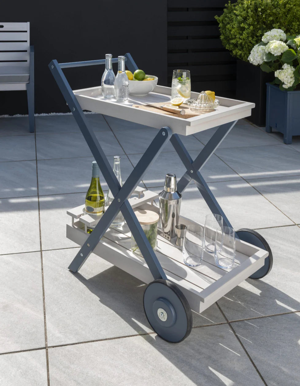 A stylish greywash drinks trolley with glass holders and wheels, set on a patio with outdoor furniture in the background.
