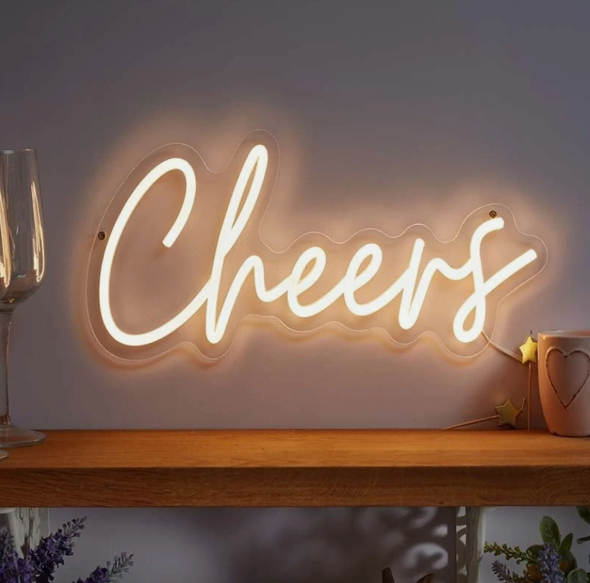 Neon 'Cheers' sign above a bar setup with drinks and bottles on a wooden deck.