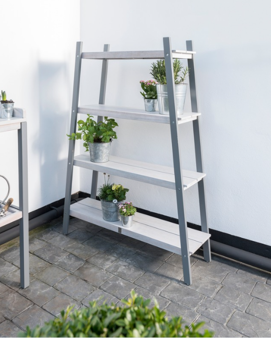 A greywash-finished Grigio Plant Shelf with multiple tiers, displaying various plants and pots, placed outdoors on a paved area.