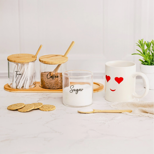 Set of glass containers labeled 'Tea', 'Coffee', and 'Sugar' with a white mug featuring heart designs on a light surface.