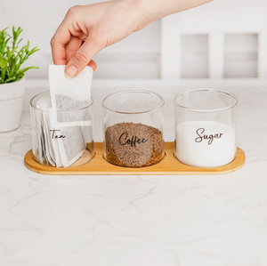 Three glass containers labeled 'Tea', 'Coffee', and 'Sugar' on a wooden tray with a hand reaching for a tea bag.