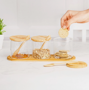 Glass jars with wooden lids and a hand reaching for a cookie on a white surface.