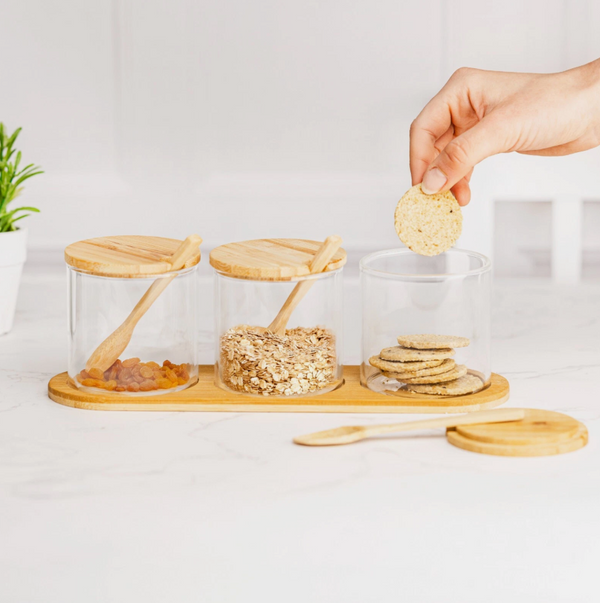 Glass jars with wooden lids and a hand reaching for a cookie on a white surface.