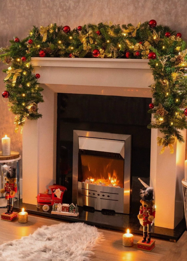 Christmas wreath with a gold bow and red ornaments layed upon a lit fire place on a warm-toned background.