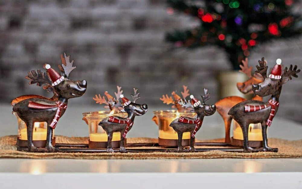 A set of metal reindeer tealight candle holders with Santa hats and scarves, holding lit candles, against a festive Christmas decorations background.