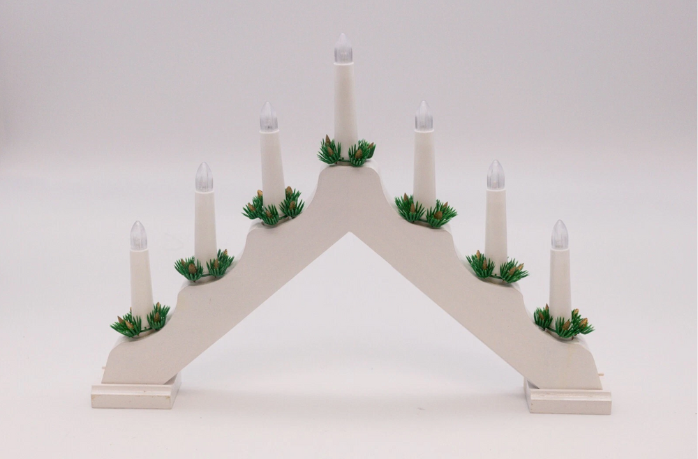 A white wooden Christmas candle bridge with seven bulbs, decorated with greenery at the base of each candle, displayed against a festive bokeh background.
