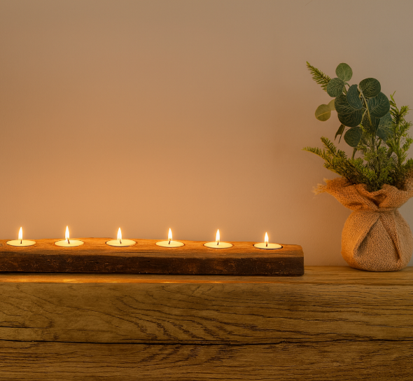 Row of candles on a wooden holder with a small plant in the background on a wooden surface.