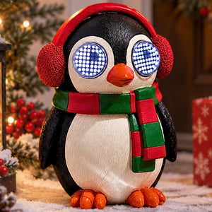 A penguin-shaped decorative figure wearing a red ear warmer hat and a red and green striped scarf, with glowing blue LED-style eyes and orange feet, set in a festive holiday scene with lights and a wrapped gift in the background.