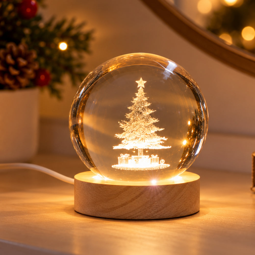 A glowing Christmas tree snow globe lamp with a warm light inside a clear sphere, set on a round wooden base on a tabletop. A blurred holiday plant appears in the background.